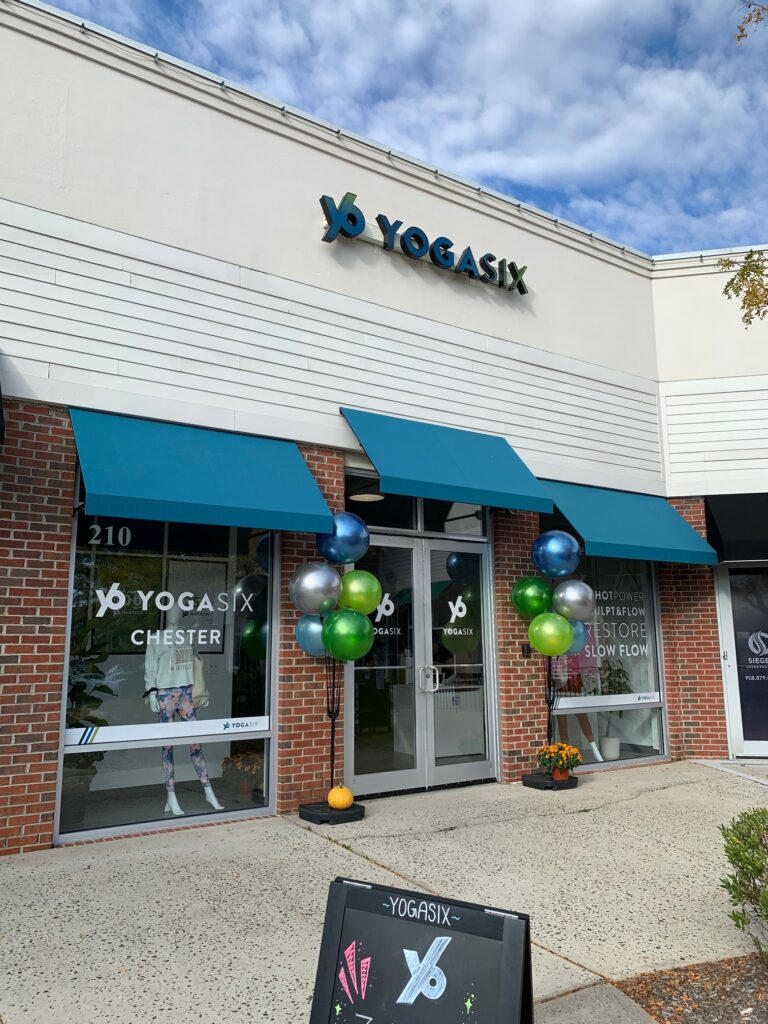 Modern retail awning installation at YogaSix Chester NJ, with fabric window canopies and branded storefront glass for yoga studio. Front view of YogaSix Chester with new Sunbrella® commercial awnings, balloons, and chalkboard sign outside yoga studio.