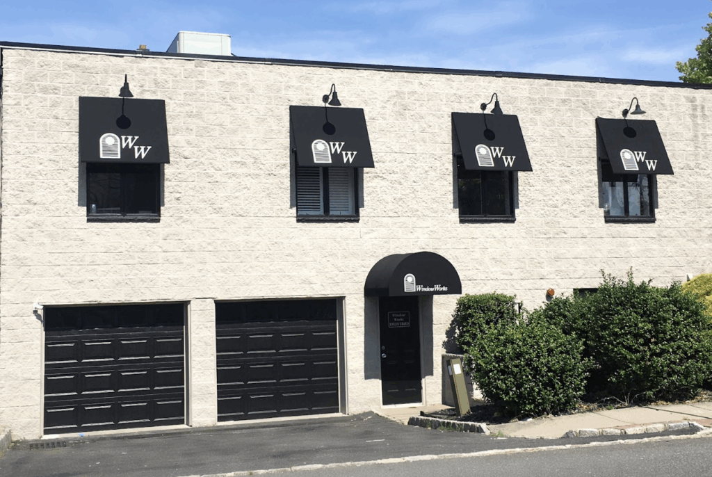 Front exterior of Window Works New Jersey headquarters with custom black branded awnings over windows and entrance, matching commercial fabric awnings with company logo and garage bay doors below.